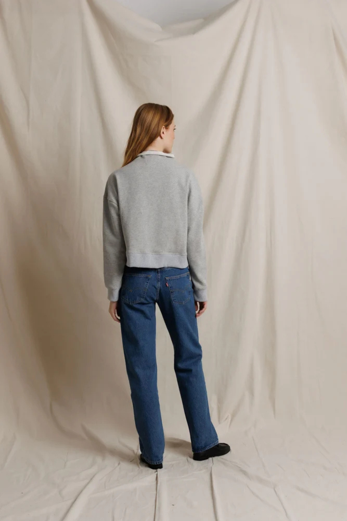 A woman stands with her back to the camera wearing a light gray cropped sweater and blue jeans, standing against a draped beige fabric backdrop.