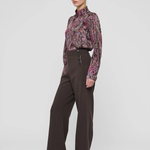 A model stands in a studio wearing a long-sleeve high-neck blouse with a purple and pink paisley pattern, paired with dark brown wide-leg trousers and black pointed-toe heels, against a light gray background.