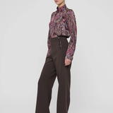 A model stands in a studio wearing a long-sleeve high-neck blouse with a purple and pink paisley pattern, paired with dark brown wide-leg trousers and black pointed-toe heels, against a light gray background.