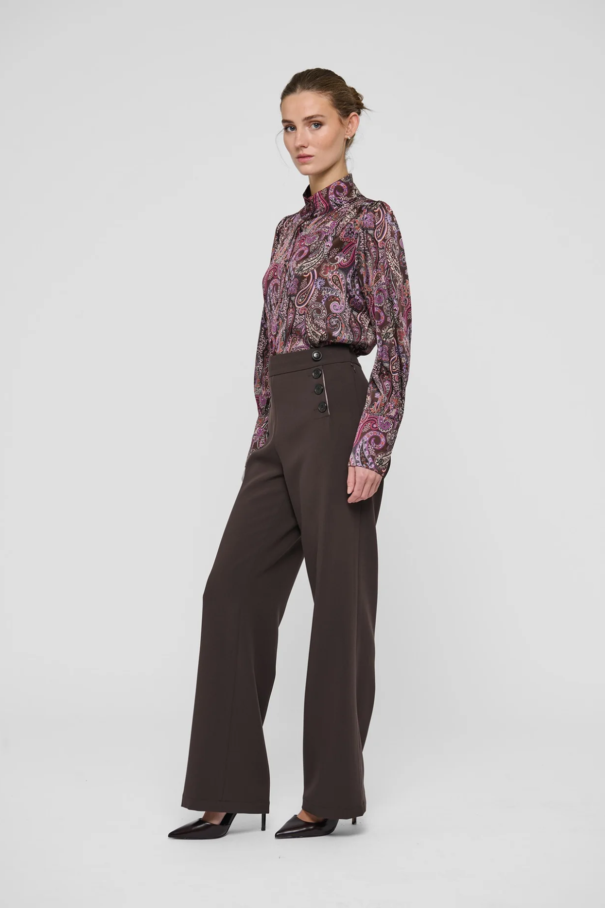 A model stands in a studio wearing a long-sleeve high-neck blouse with a purple and pink paisley pattern, paired with dark brown wide-leg trousers and black pointed-toe heels, against a light gray background.