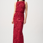 A woman modeling a sleeveless, floor-length red gown with tonal floral lace applique, photographed against a plain light gray studio background.