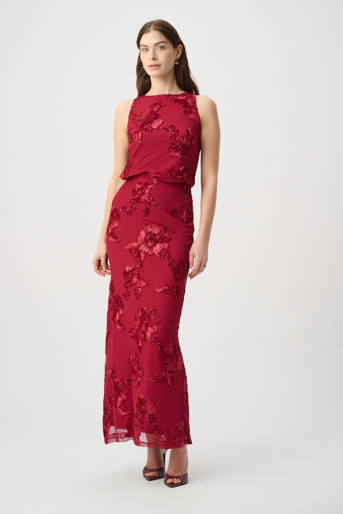A woman modeling a sleeveless, floor-length red gown with tonal floral lace applique, photographed against a plain light gray studio background.