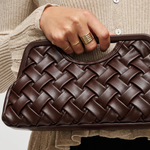 A close-up of a person holding a dark brown woven leather clutch bag. The person is wearing a beige ribbed cardigan and black bottoms, with gold rings on their fingers.