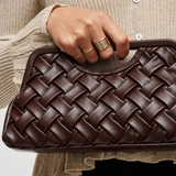 A close-up of a person holding a dark brown woven leather clutch bag. The person is wearing a beige ribbed cardigan and black bottoms, with gold rings on their fingers.