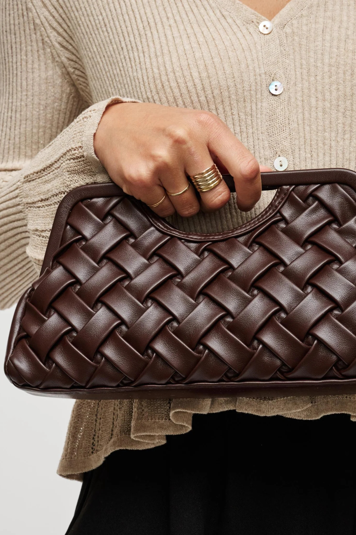 A close-up of a person holding a dark brown woven leather clutch bag. The person is wearing a beige ribbed cardigan and black bottoms, with gold rings on their fingers.