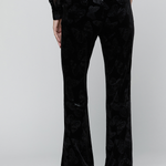 Black velvet flare pants with a tonal floral jacquard pattern, shown on a model from the back against a light gray studio background.