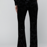 Black velvet flare pants with a tonal floral jacquard pattern, shown on a model from the back against a light gray studio background.