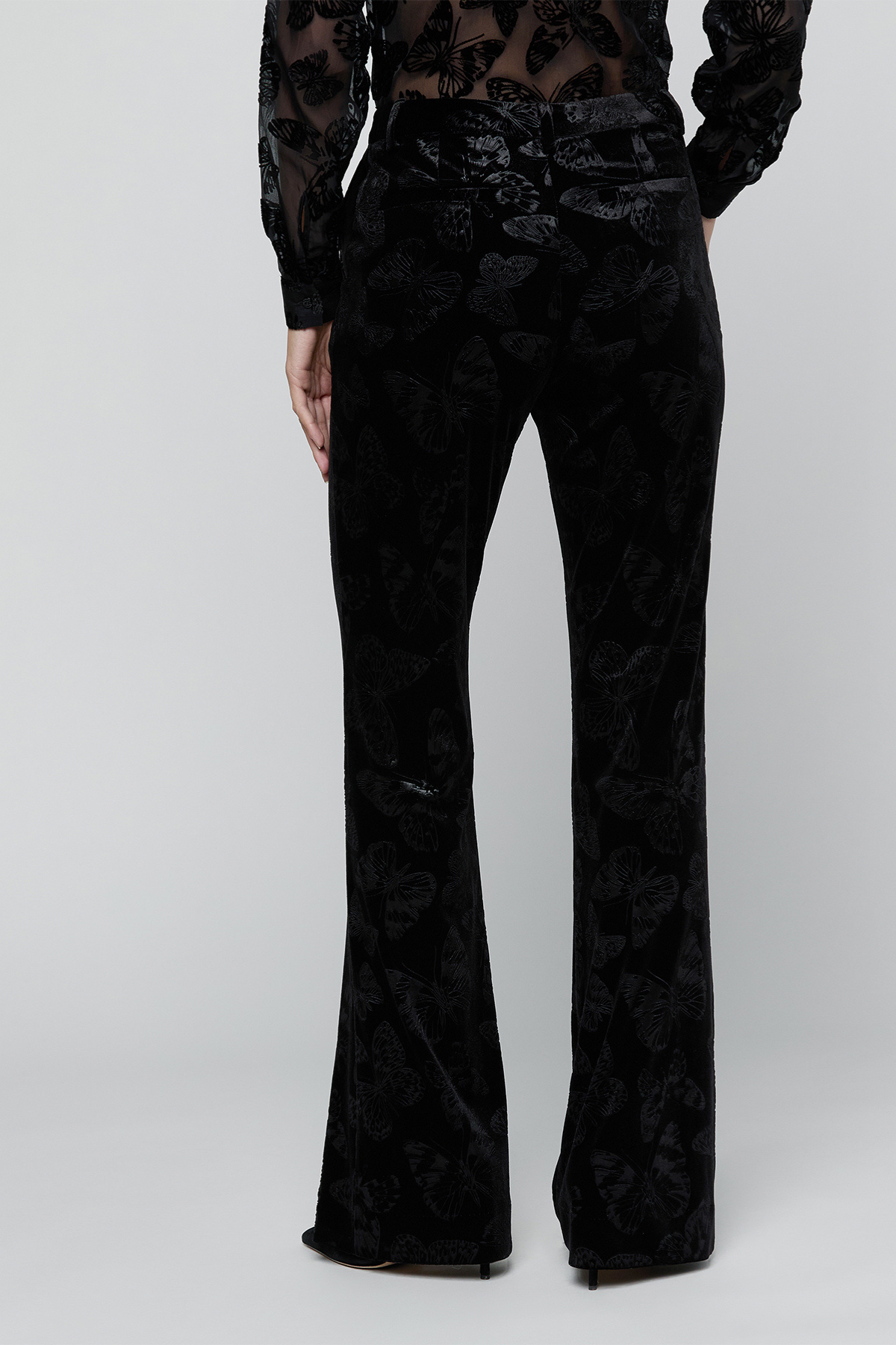 Black velvet flare pants with a tonal floral jacquard pattern, shown on a model from the back against a light gray studio background.