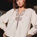 A woman wearing a cream-colored embroidered blouse with long puff sleeves and a decorative embroidered yoke, paired with olive green pants, in a dimly lit indoor setting.
