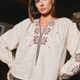 A woman wearing a cream-colored embroidered blouse with long puff sleeves and a decorative embroidered yoke, paired with olive green pants, in a dimly lit indoor setting.