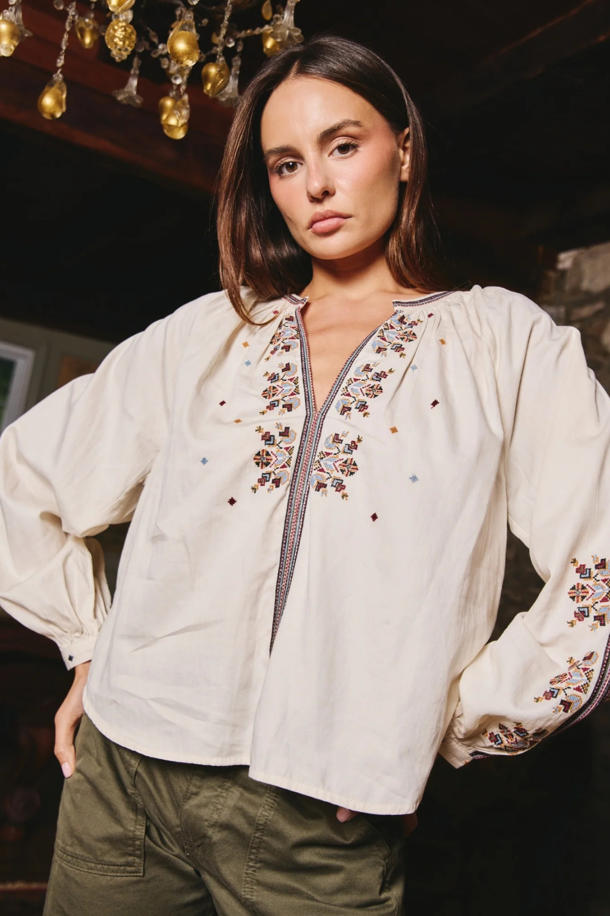 A woman wearing a cream-colored embroidered blouse with long puff sleeves and a decorative embroidered yoke, paired with olive green pants, in a dimly lit indoor setting.