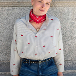 A woman wearing a cream-colored long-sleeve button-up shirt with red cherry prints, a red bandana, and blue jeans, standing against a gray stone wall.
