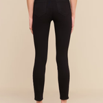 Rear view of a model wearing black skinny jeans with a textured black belt at the waist, paired with a white top and black heels, against a solid beige background.
