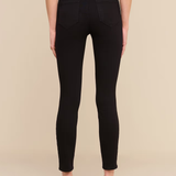 Rear view of a model wearing black skinny jeans with a textured black belt at the waist, paired with a white top and black heels, against a solid beige background.