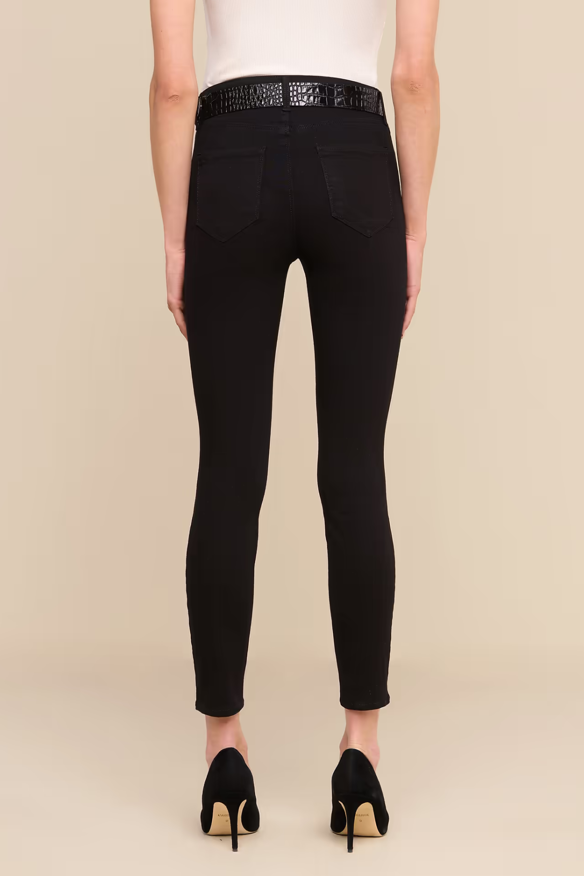 Rear view of a model wearing black skinny jeans with a textured black belt at the waist, paired with a white top and black heels, against a solid beige background.