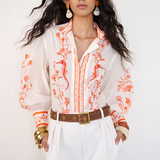 Woman wearing a white blouse with orange floral patterns and white pants against a white background
