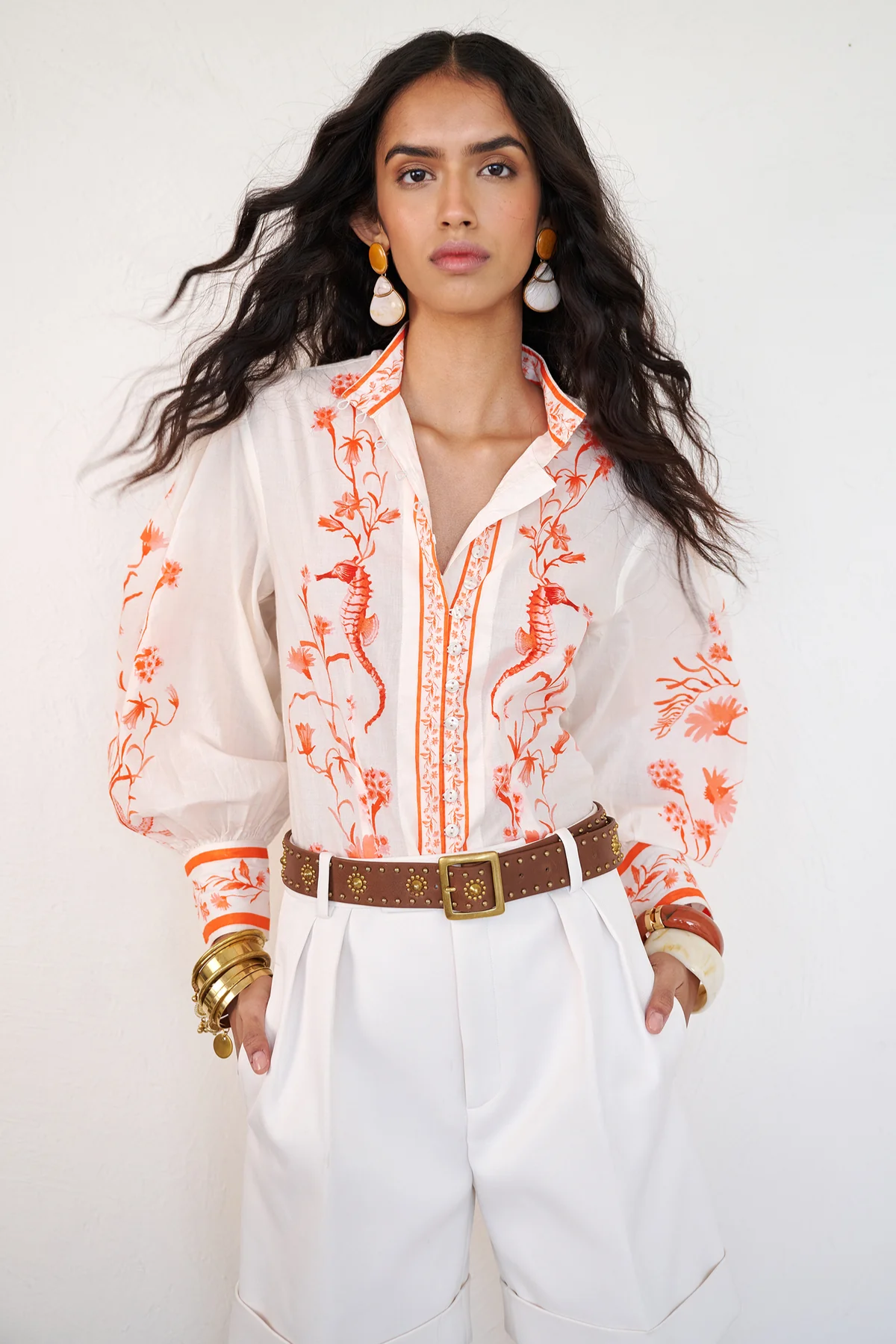 Woman wearing a white blouse with orange floral patterns and white pants against a white background