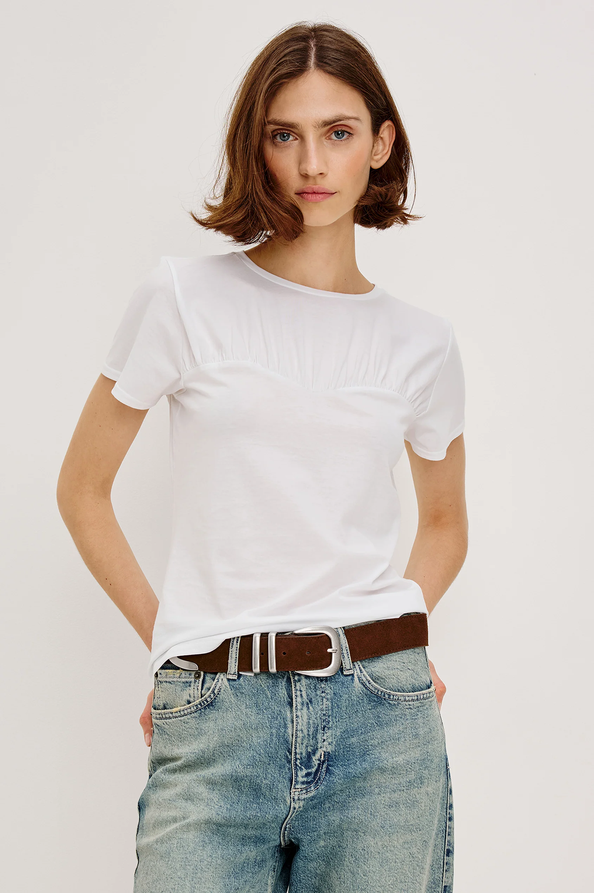 Person wearing a white t-shirt and blue jeans with a brown belt on a plain background