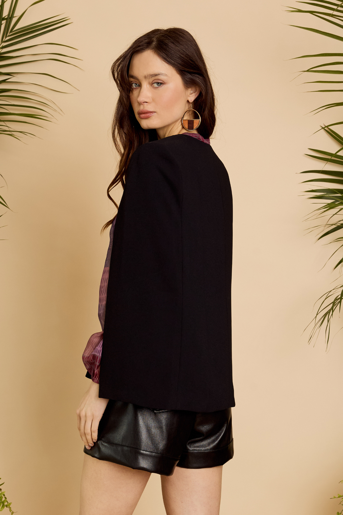 Woman wearing a black jacket and leather shorts against a beige background with green foliage.