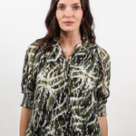 A woman wearing a green, black, and white abstract patterned blouse with three-quarter sleeves and blue jeans, standing against a plain light gray background.