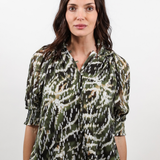 A woman wearing a green, black, and white abstract patterned blouse with three-quarter sleeves and blue jeans, standing against a plain light gray background.