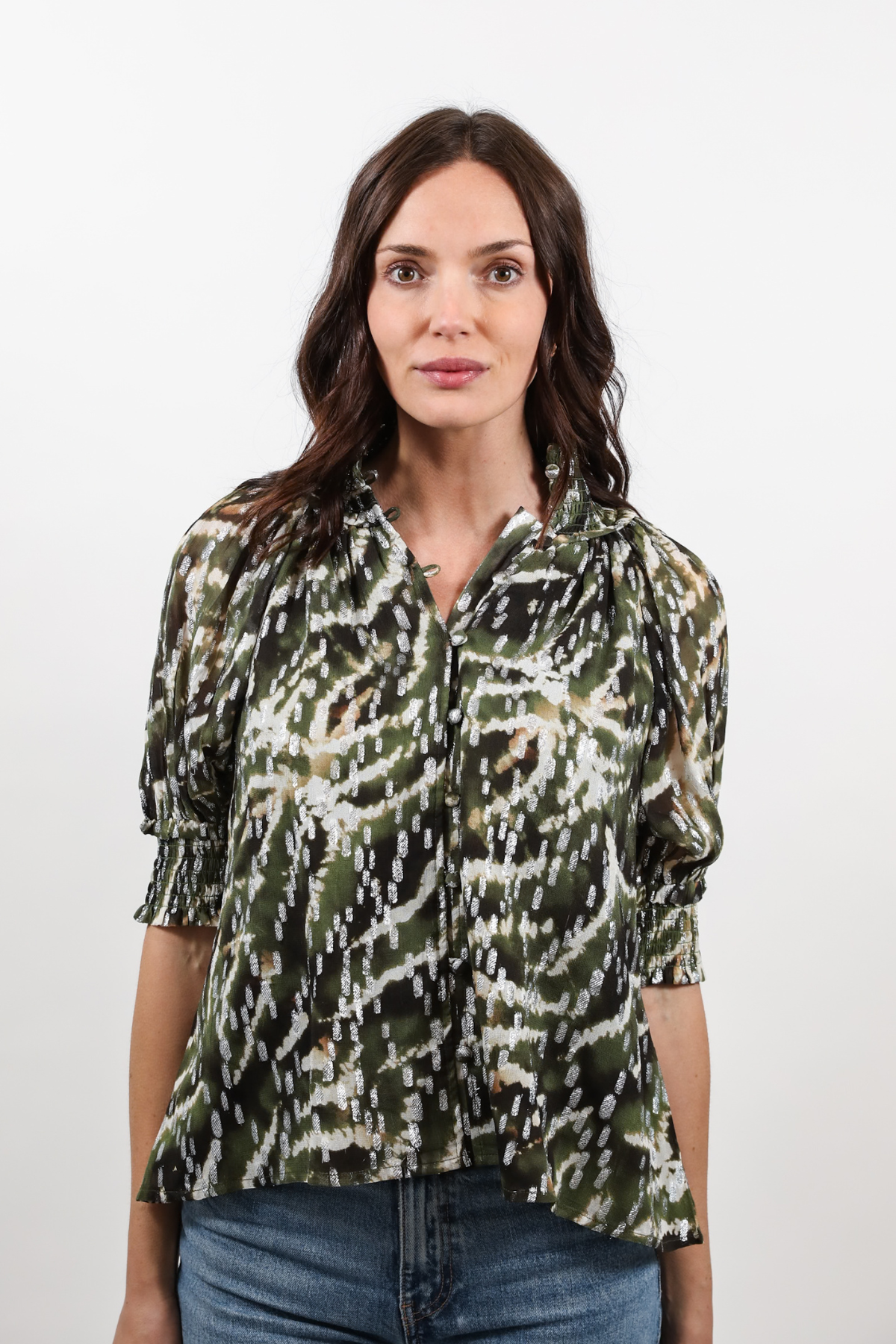 A woman wearing a green, black, and white abstract patterned blouse with three-quarter sleeves and blue jeans, standing against a plain light gray background.