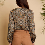 A woman viewed from the back wearing a long-sleeve blouse with an intricate geometric and tribal-style pattern in brown, beige, black, and muted orange, paired with tan pants, set against a warm beige background with decorative palm leaves.