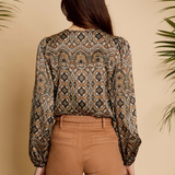 A woman viewed from the back wearing a long-sleeve blouse with an intricate geometric and tribal-style pattern in brown, beige, black, and muted orange, paired with tan pants, set against a warm beige background with decorative palm leaves.