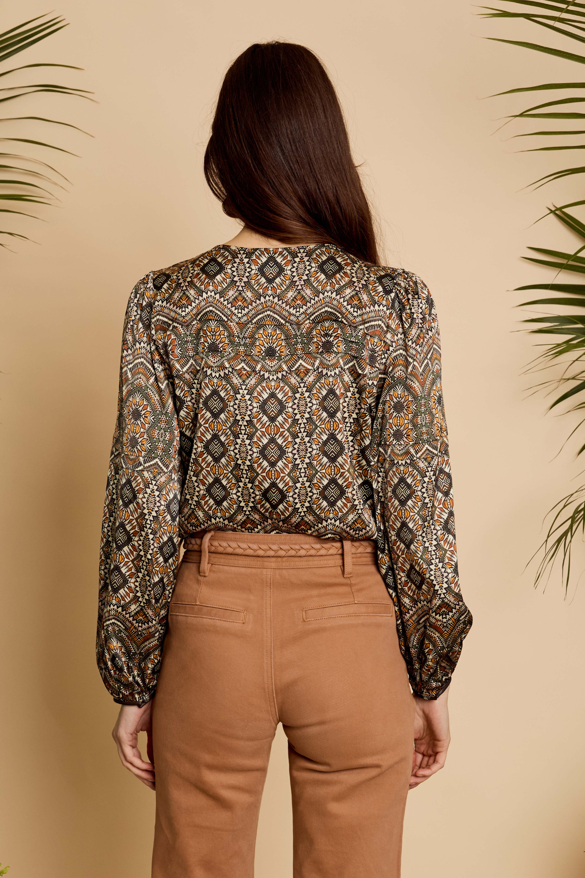 A woman viewed from the back wearing a long-sleeve blouse with an intricate geometric and tribal-style pattern in brown, beige, black, and muted orange, paired with tan pants, set against a warm beige background with decorative palm leaves.