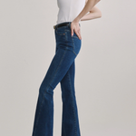 A model wearing a pair of dark blue high-waisted flare jeans with a black belt, paired with a white sleeveless top and black shoes, against a light gray studio background.