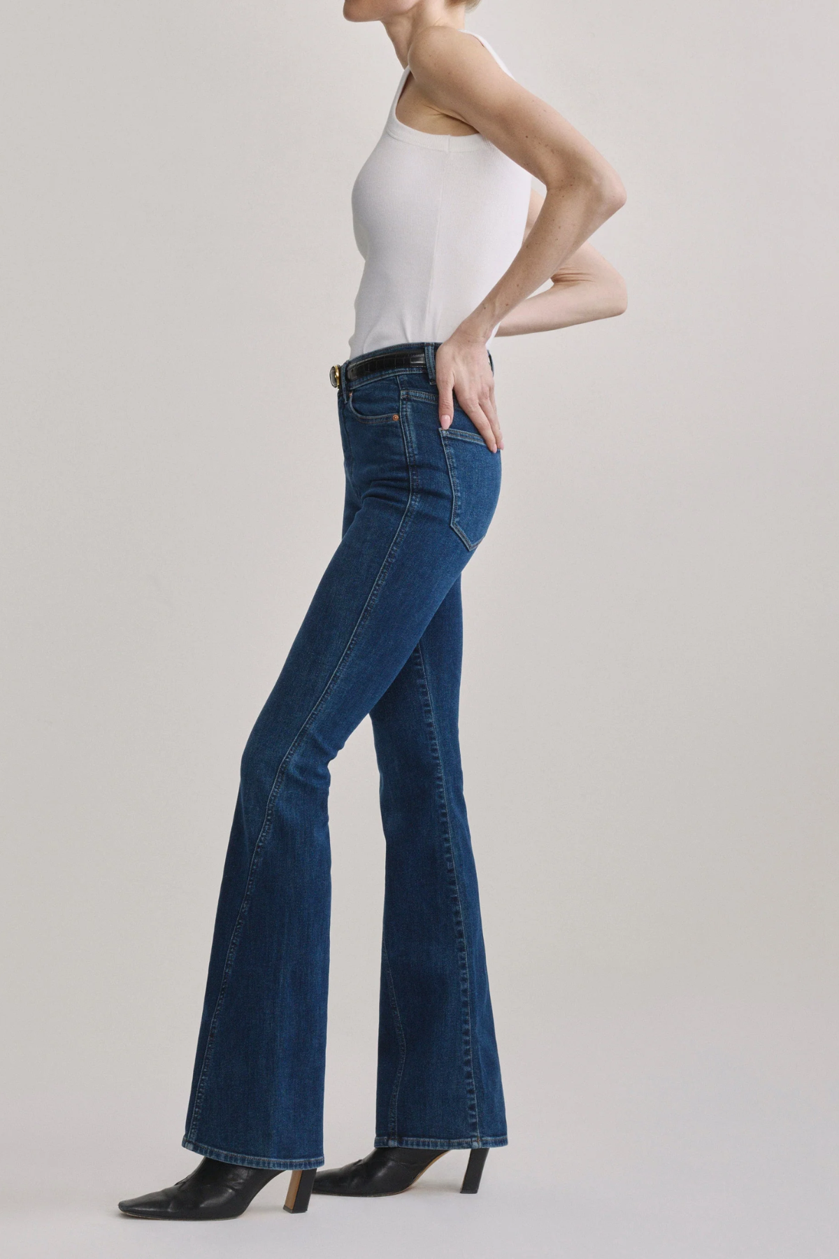 A model wearing a pair of dark blue high-waisted flare jeans with a black belt, paired with a white sleeveless top and black shoes, against a light gray studio background.