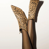 A pair of leopard-print ankle boots worn with sheer black tights, shown against a plain off-white background with a strong shadow.
