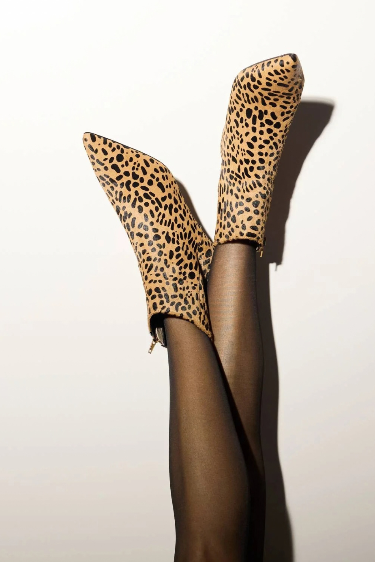 A pair of leopard-print ankle boots worn with sheer black tights, shown against a plain off-white background with a strong shadow.