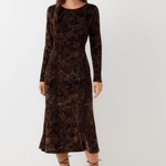 A woman modeling a long-sleeve midi dress in dark brown velvet with an intricate floral jacquard pattern, paired with black lace-up strappy heels, against a plain light gray studio background.