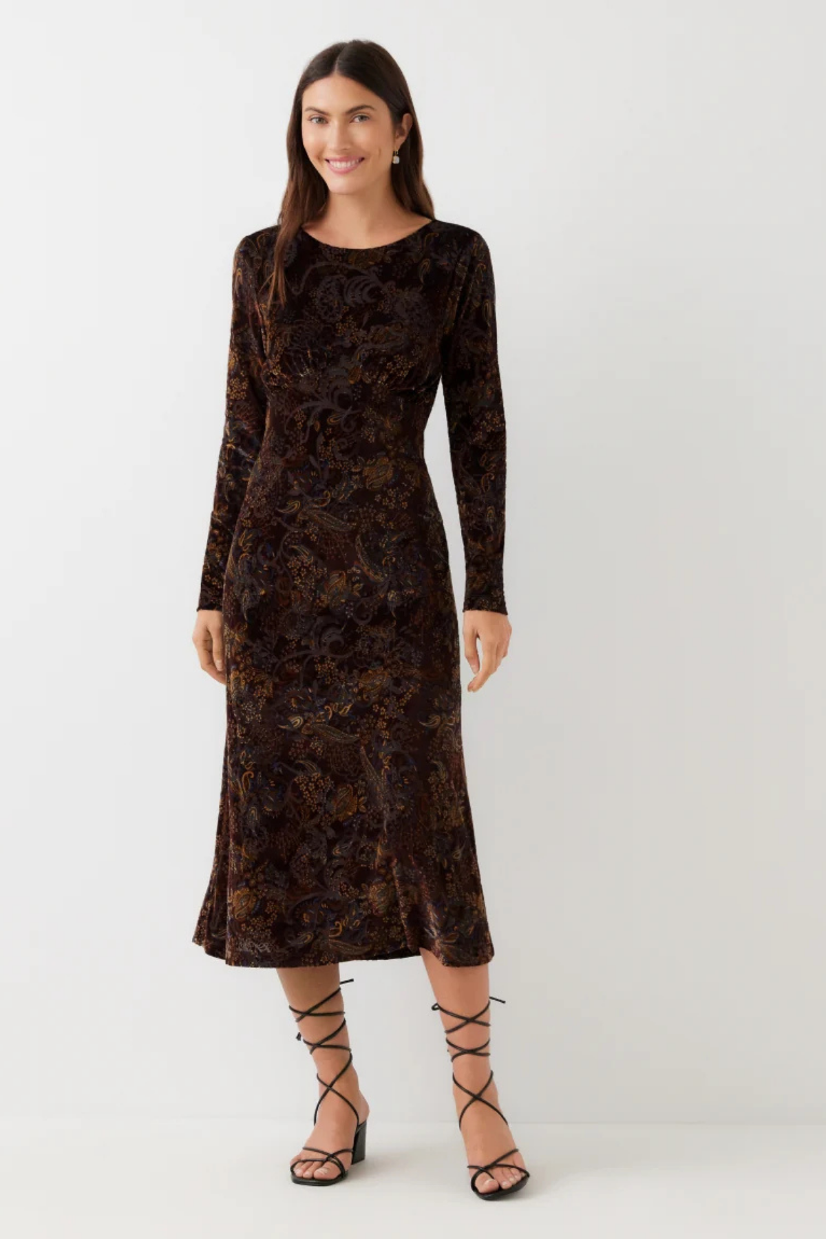 A woman modeling a long-sleeve midi dress in dark brown velvet with an intricate floral jacquard pattern, paired with black lace-up strappy heels, against a plain light gray studio background.