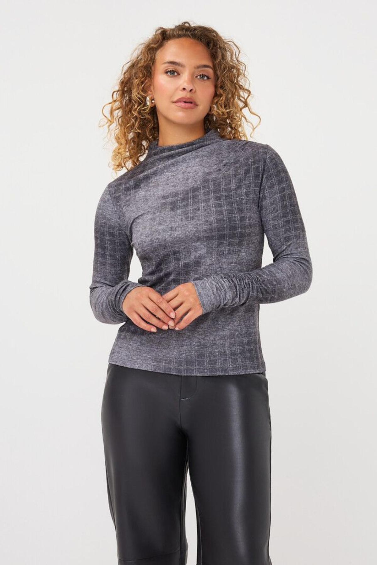 A woman with curly light brown hair wearing a gray textured long-sleeve mock neck top and black faux leather pants, standing against a light gray studio background.