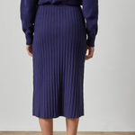 A person shown from the back wearing a dark purple ribbed knit midi skirt with a matching long-sleeve top and brown leather loafers, set against a plain light gray wall and beige floor.