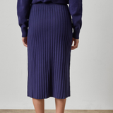A person shown from the back wearing a dark purple ribbed knit midi skirt with a matching long-sleeve top and brown leather loafers, set against a plain light gray wall and beige floor.