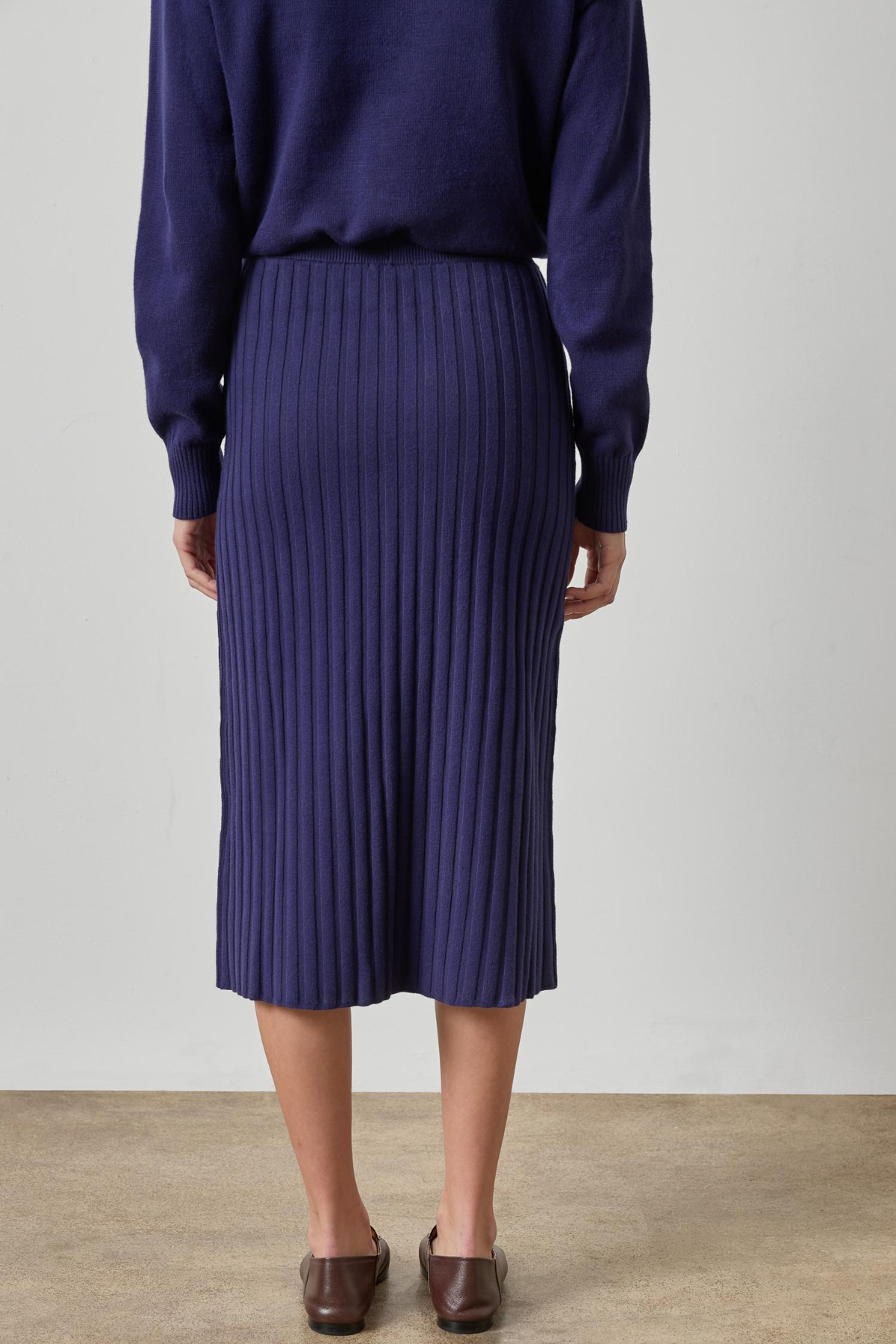 A person shown from the back wearing a dark purple ribbed knit midi skirt with a matching long-sleeve top and brown leather loafers, set against a plain light gray wall and beige floor.