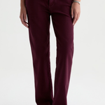 A person wearing straight-leg pants in a deep burgundy or plum color, paired with brown suede loafers and a brown top, photographed against a clean white studio background.