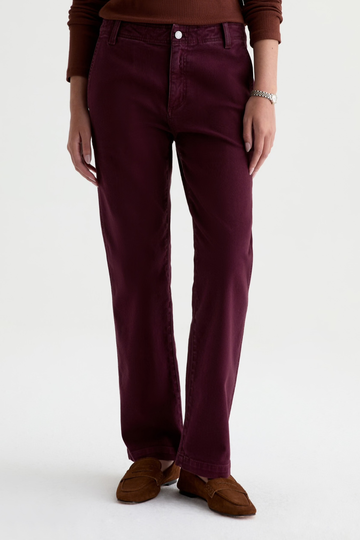 A person wearing straight-leg pants in a deep burgundy or plum color, paired with brown suede loafers and a brown top, photographed against a clean white studio background.