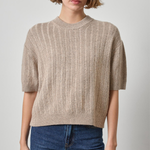 A woman wearing a beige short-sleeve knit sweater with vertical ribbing and blue jeans, standing against a light gray studio background.