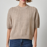 A woman wearing a beige short-sleeve knit sweater with vertical ribbing and blue jeans, standing against a light gray studio background.