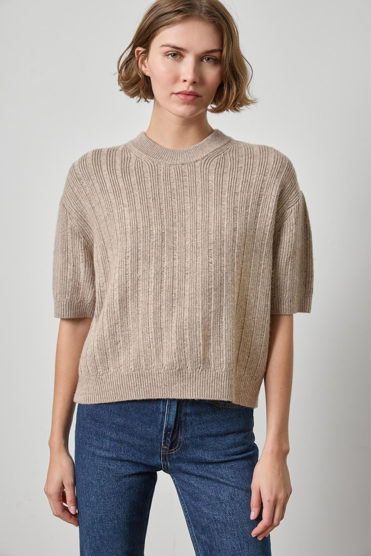 A woman wearing a beige short-sleeve knit sweater with vertical ribbing and blue jeans, standing against a light gray studio background.