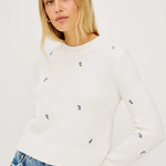 Woman wearing a white sweater with small colorful patterns and blue jeans on a plain background