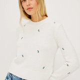 Woman wearing a white sweater with small colorful patterns and blue jeans on a plain background