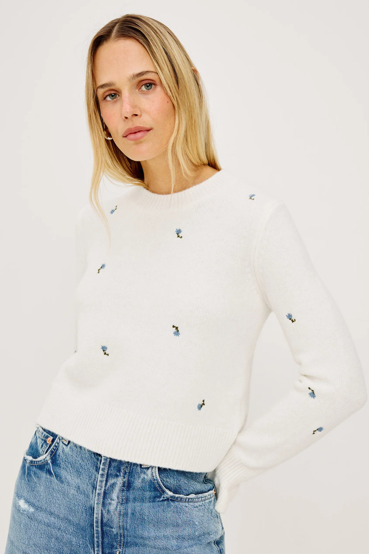 Woman wearing a white sweater with small colorful patterns and blue jeans on a plain background