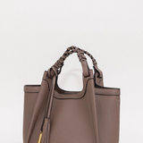 A taupe brown pebbled leather handbag with braided top handles, a tassel detail, and gold hardware, shown on a plain light gray background.