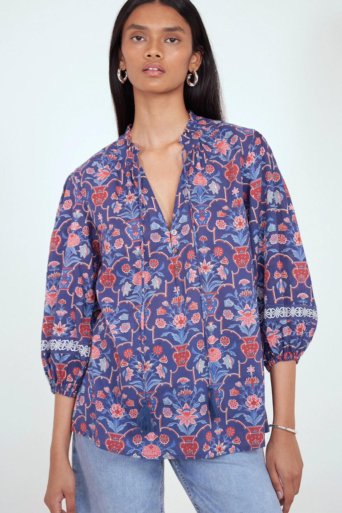 A woman wearing a blue floral print blouse with red and pink floral motifs, paired with light blue jeans, against a plain white studio background.