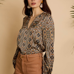 A woman modeling a long-sleeve V-neck blouse with a brown, beige, and black geometric pattern, paired with brown pants, set against a warm beige background with decorative palm leaves.